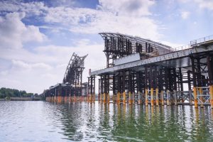 Steel framework of a large bridge under construction across a river on a sunny day.